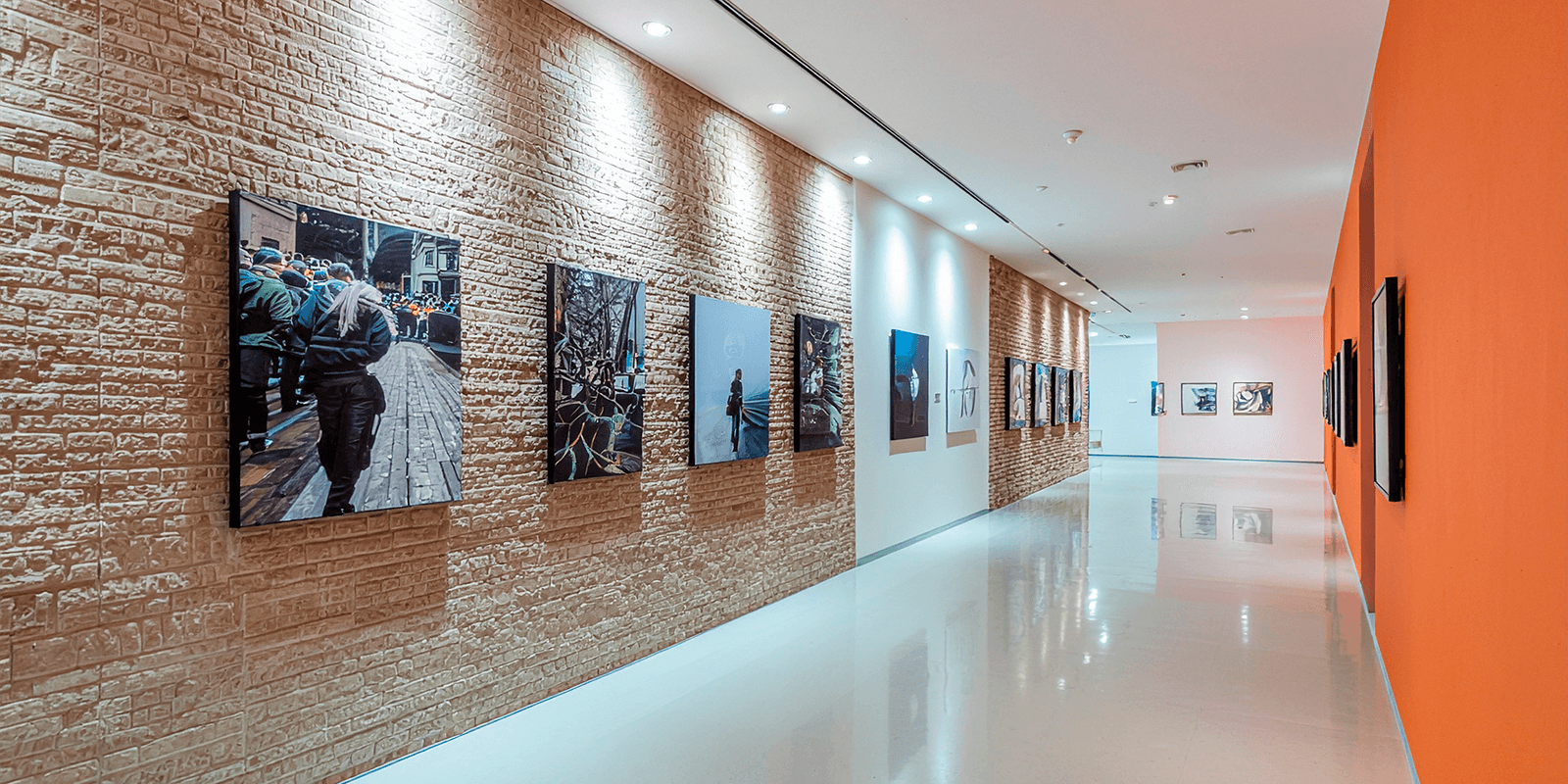 Gallery corridor with framed photographs on exposed brick and orange accent wall, spotlit exhibition panels