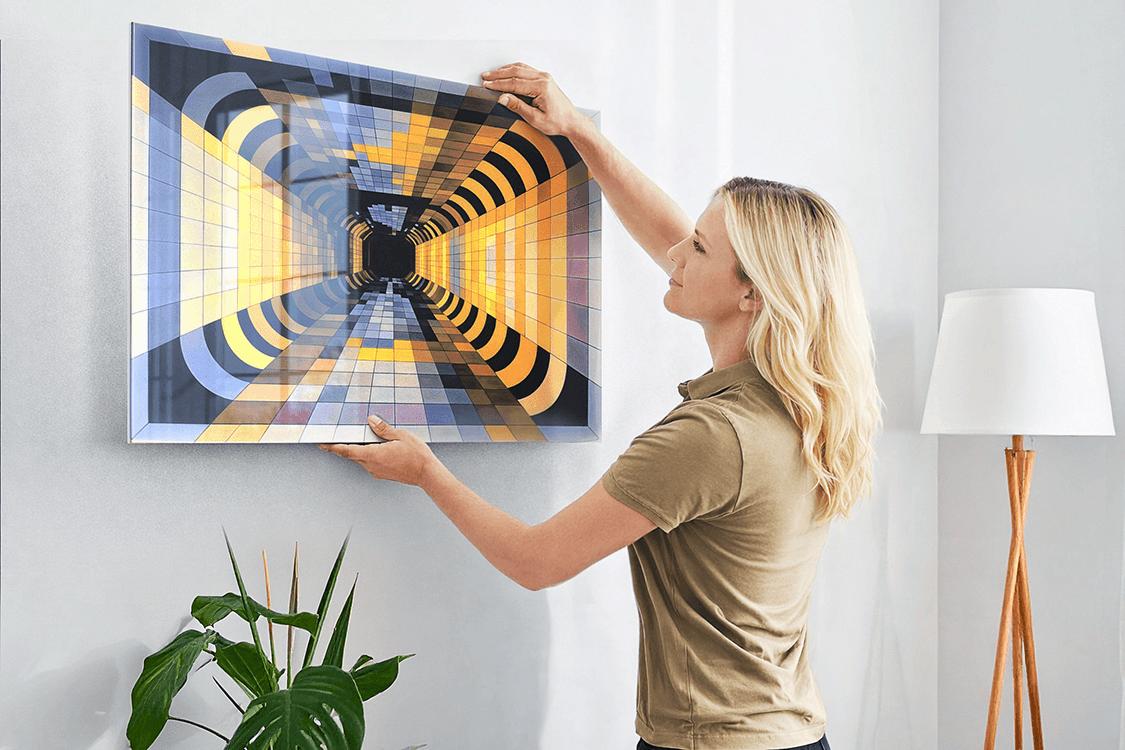 Woman with long blonde hair in an olive top hanging a large Acrylux Gallery print with a geometric tunnel artwork on a white living-room wall