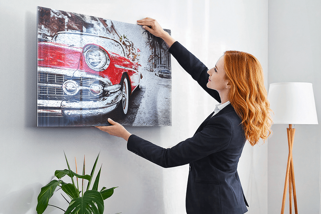 Red-haired woman in a dark blazer hanging an Acrylux Metallic print of a vintage red car on a monochrome street against a white living-room wall