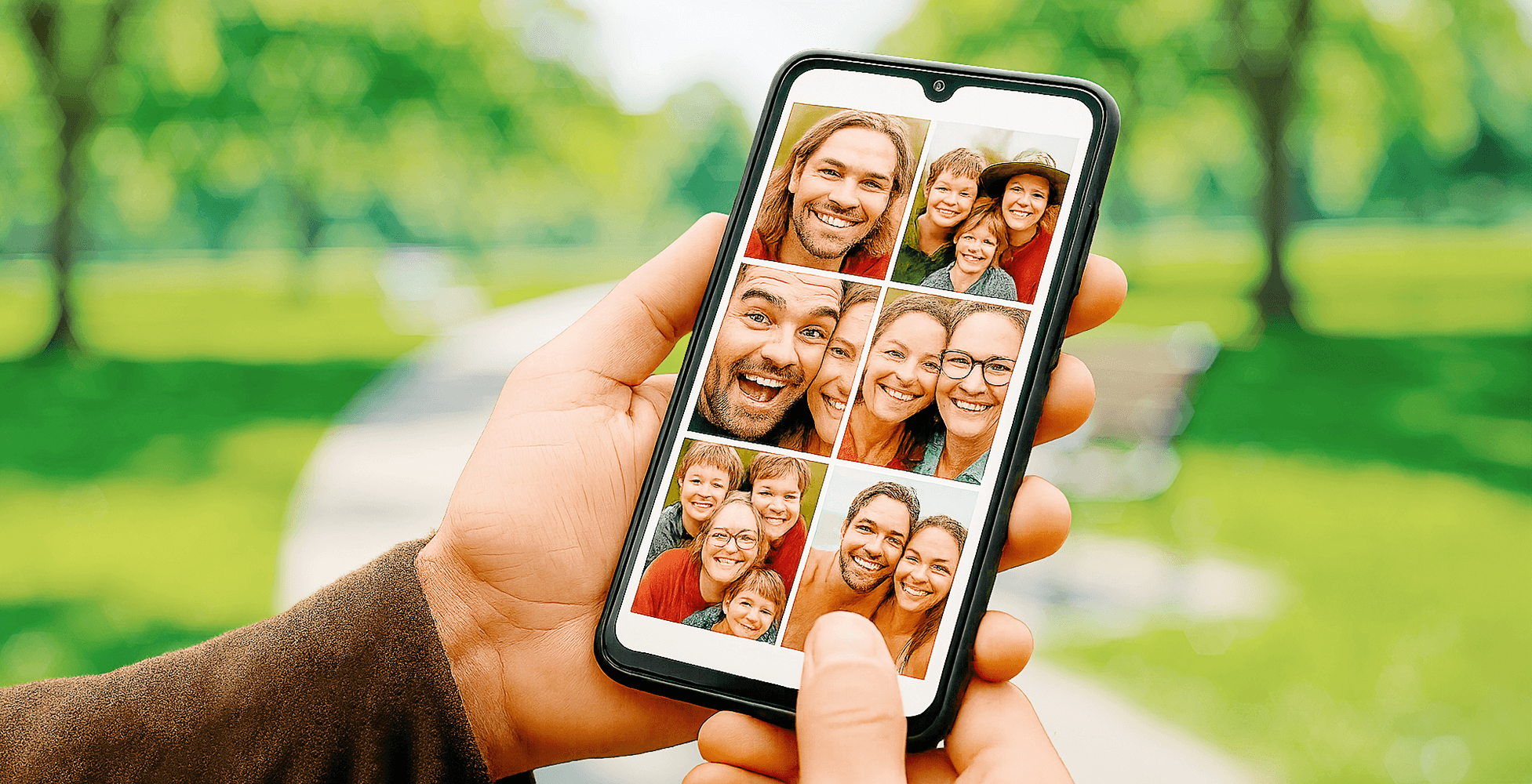 Hand holding smartphone with photo album open, in a park, showing grid of family photos